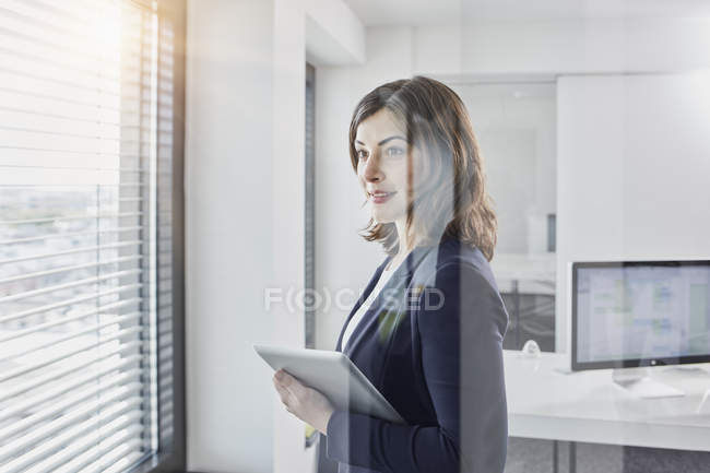 Joven empresaria sonriente con tableta mirando por la ventana en la oficina - foto de stock
