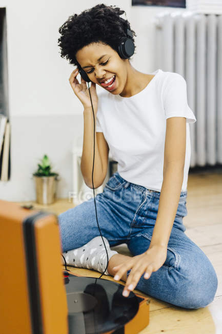Young african american woman sitting on ground and listening music from record player, using headphones — Stock Photo