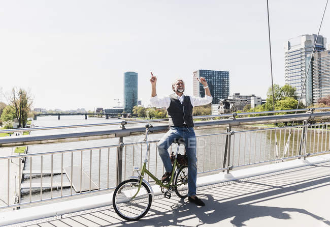 Happy mature man with bicycle listening to music on bridge in the city — Stock Photo