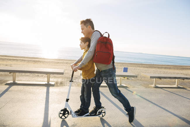 Vater und Sohn fahren Roller auf Strandpromenade bei Sonnenuntergang — Stockfoto