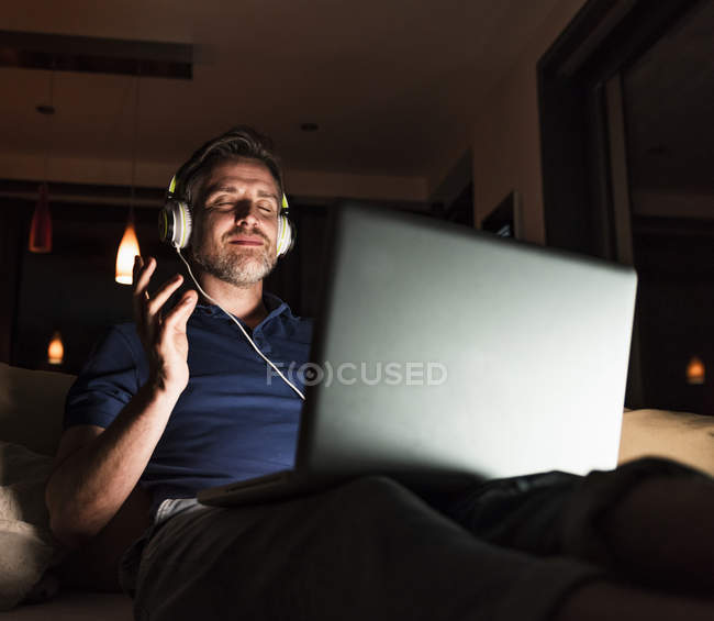 Homme assis sur le canapé à la maison écoutant de la musique avec écouteurs et ordinateur portable — Photo de stock