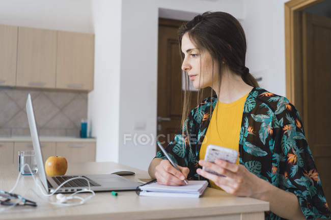 Молодая женщина сидит за столом дома с помощью мобильного телефона и ноутбука, делая заметки — стоковое фото