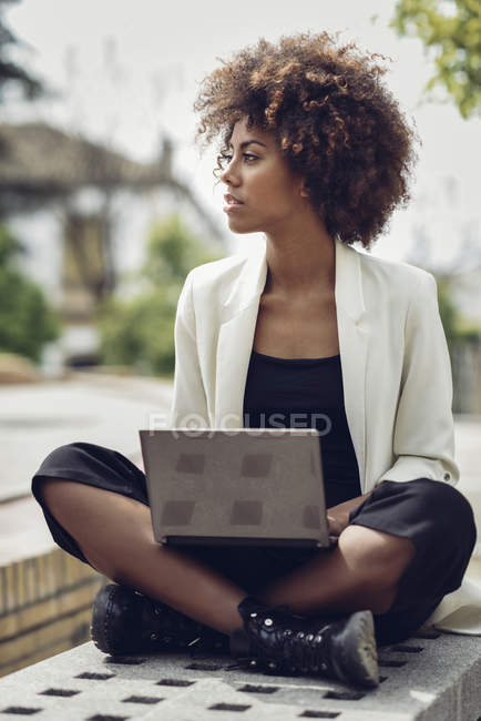 Fashionable young woman with curly hair sitting on bench with laptop — Stock Photo