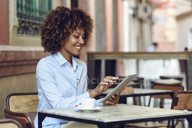 Femme souriante avec coiffure afro assis dans un café en plein air en utilisant une tablette — Photo de stock