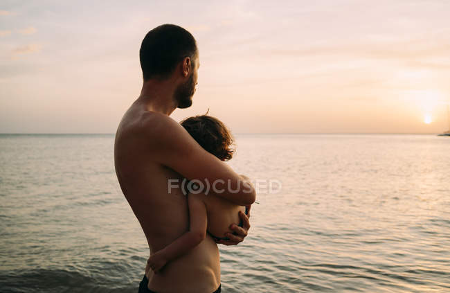 Батько тримає маленьку дочку на руках, спостерігаючи захід сонця разом у морі. — Stock Photo
