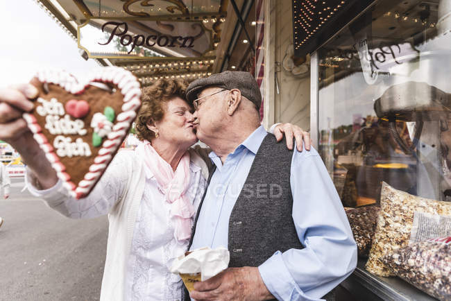 Старшая пара с пряничным сердечком целуется на ярмарке — стоковое фото