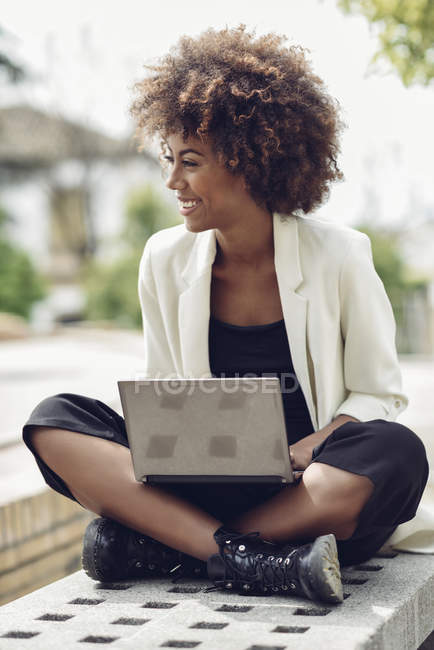Модна молода жінка з Кучеряве волосся сидячи на лавці з ноутбуком сміятися — стокове фото