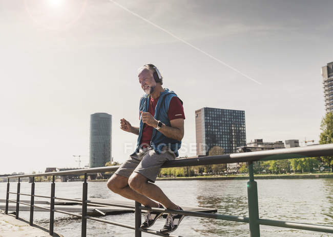 Feliz atlético maduro homem com fones de ouvido sentado à beira do rio na cidade — Fotografia de Stock