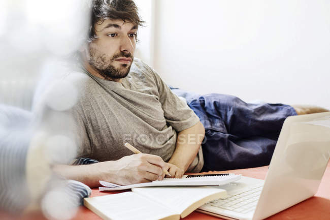 Porträt eines Studenten, der mit Laptop und Buch im Bett liegt — Stockfoto