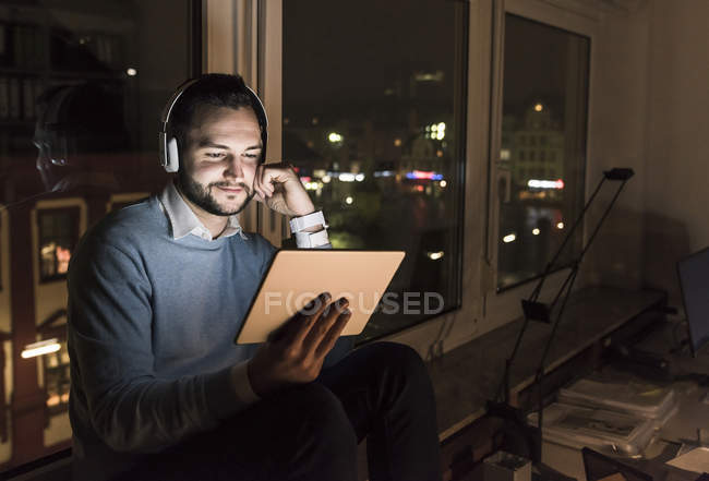 Бизнесмен, сидящий ночью на подоконнике в офисе с планшетами и наушниками — стоковое фото