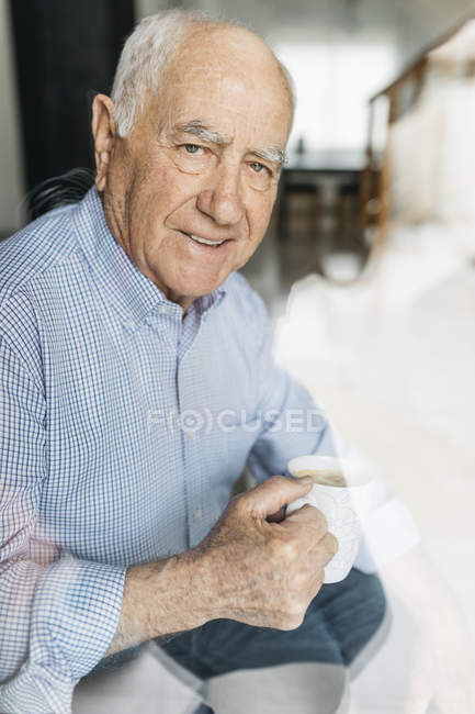 Hombre mayor tomando un café mientras se relaja en casa, mirando a la cámara a través de un vaso - foto de stock