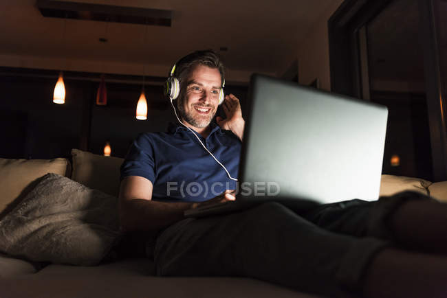 Человек в наушниках сидит дома на диване и смотрит на ноутбук. — стоковое фото