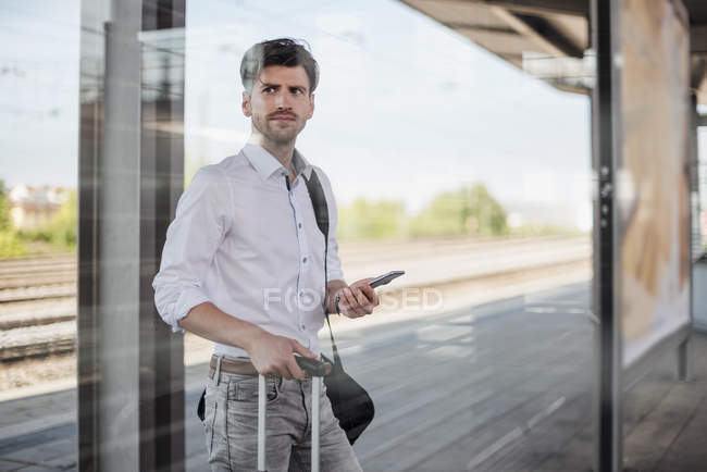 Geschäftsmann mit Gepäck und Handy steht auf Bahnsteig und schaut sich um — Stockfoto