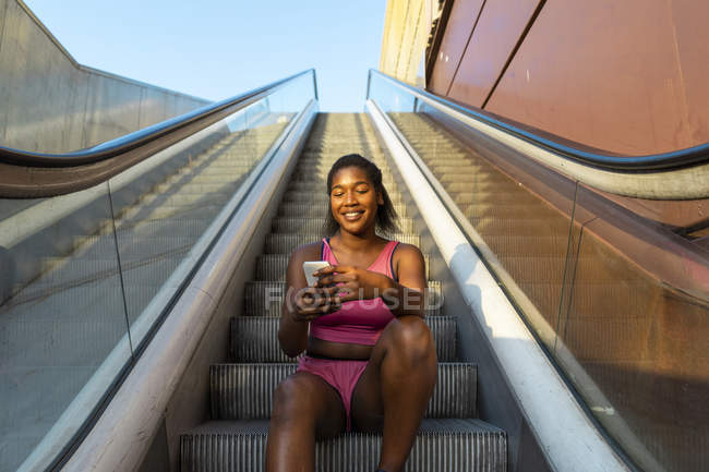 Young woman sitting on escalator, using smartphone — Stock Photo