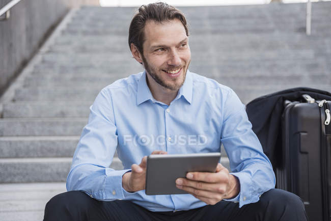 Uomo d'affari sorridente con valigia rotolante seduto sulle scale utilizzando tablet — Foto stock