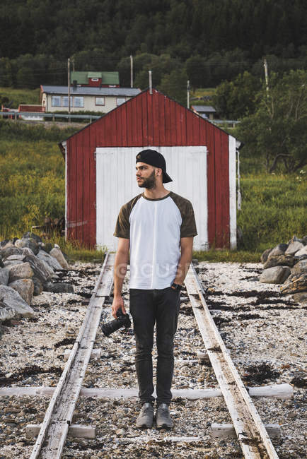 Jovem com câmera em pé na frente de uma cabana de celeiro vermelho — Fotografia de Stock