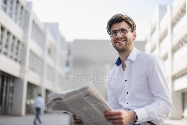 Hombre de negocios sonriente sentado en la ciudad y leyendo el periódico - foto de stock