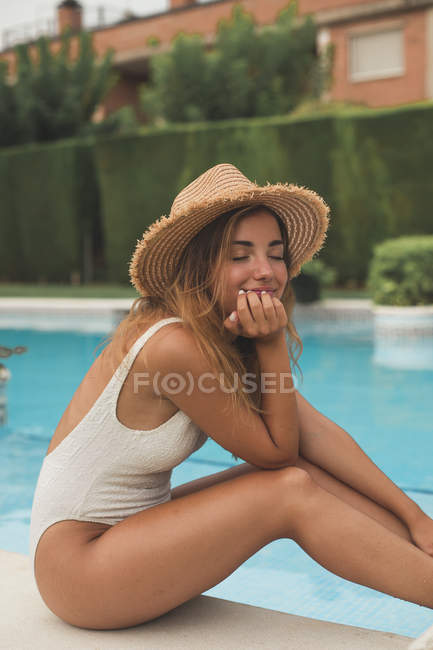 Smiling young woman with straw hat sitting at poolside with closed eyes — Stock Photo