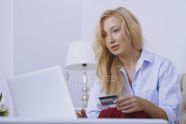 Blond woman sitting on couch, using laptop to make a payment with her credit card — Stock Photo
