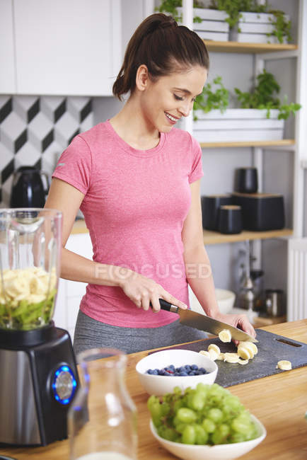 Smiling young woman preparing smoothie in kitchen, cropping banana with knife — Stock Photo
