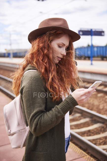 Рыжая женщина в шляпе стоит на платформе и смотрит на мобильный телефон. — стоковое фото