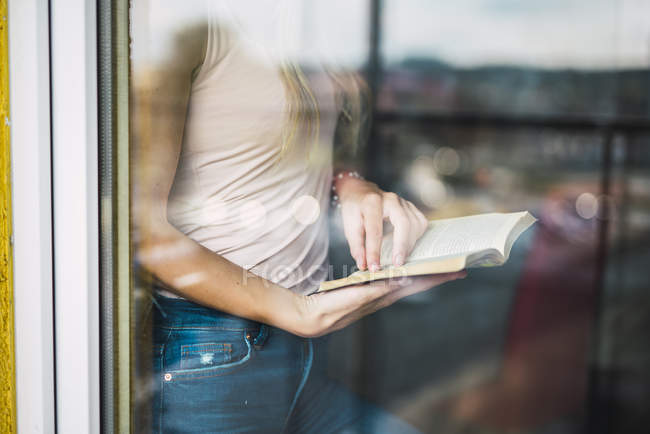 Close-up of young woman reading a book behind windowpane — Stock Photo