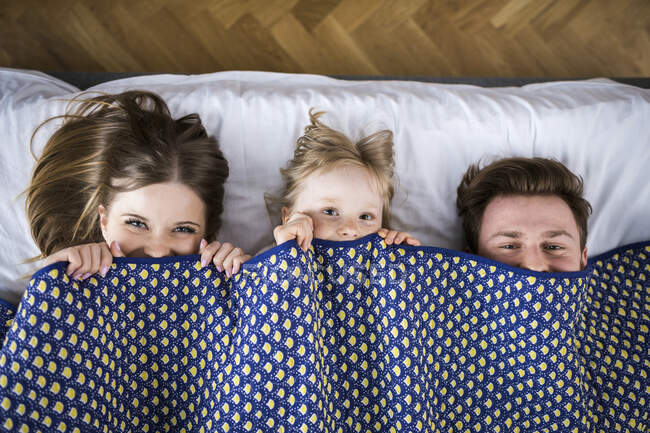 Familia feliz acostada en la cama, escondida bajo una manta - foto de stock