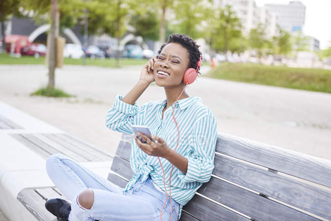 Портрет молодой женщины, сидящей на скамейке и слушающей музыку с мобильным телефоном и наушниками — стоковое фото
