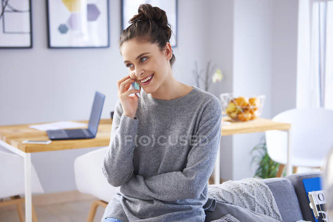 Jovem mulher sentada em casa, falando ao telefone, mesa com laptop no fundo — Fotografia de Stock