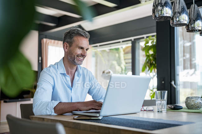 Улыбающийся взрослый мужчина дома с ноутбуком за столом — стоковое фото