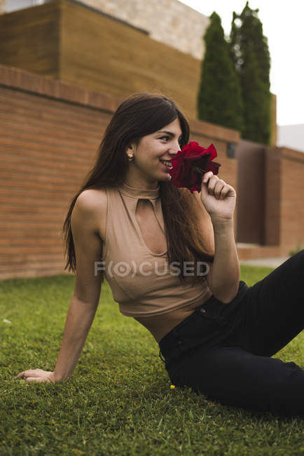Lächelnde junge Frau auf dem Rasen, die nach roter Rose riecht — Stockfoto