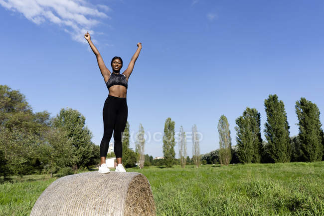 Jeune athlète debout sur une balle de paille, levant les bras sous le ciel bleu — Photo de stock