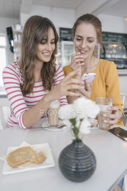 Dos novias se reúnen en una cafetería, usando un smartphone - foto de stock