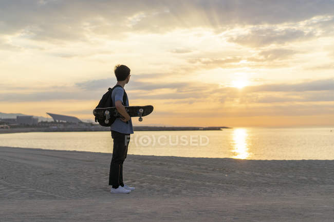 Young Chinese man with skateboard standing at the beach at sunrise — Stock Photo