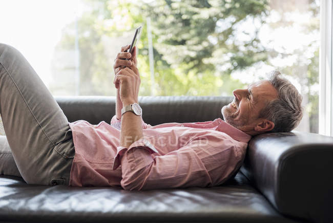 Homme mûr souriant couché sur le canapé à la maison à l'aide d'un téléphone portable — Photo de stock