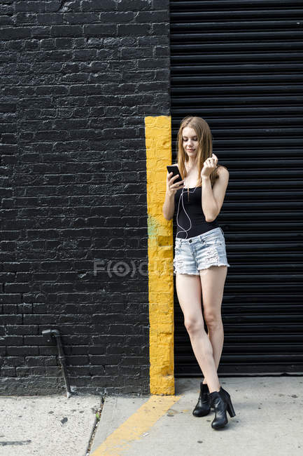 Jeune femme penchée au coin de la rue, écoutant de la musique, utilisant un smartphone — Photo de stock