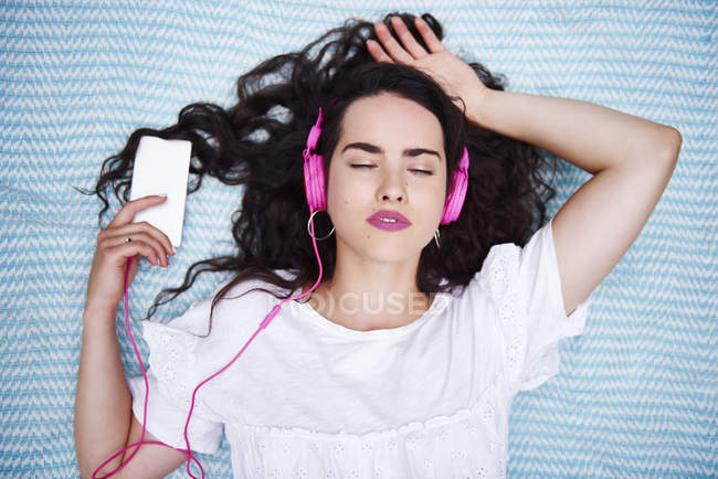 Портрет молодой женщины, лежащей на одеяле, слушающей музыку со смартфоном и розовыми наушниками — стоковое фото