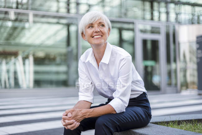 Усміхнена старша бізнес-леді сидить перед скляним фасадом і озирається навколо — стокове фото