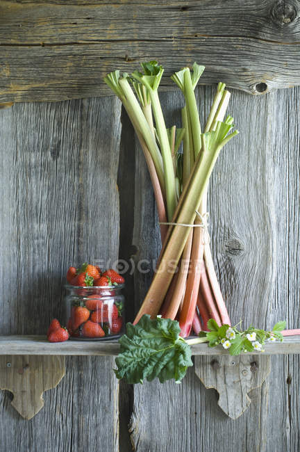 Bunch of rhubarb stalks and strawberries in a glass on rack — Stock Photo