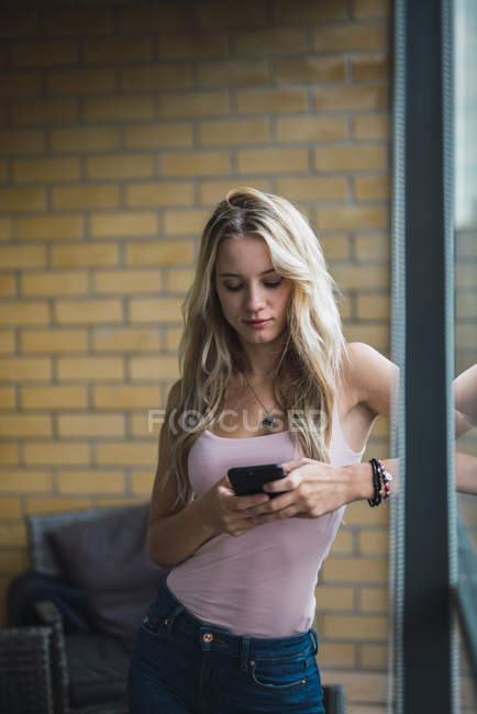 Loira jovem verificando celular na janela — Fotografia de Stock