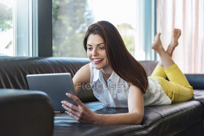 Lächelnde Frau, die zu Hause mit einem Tablet auf dem Sofa liegt — Stockfoto