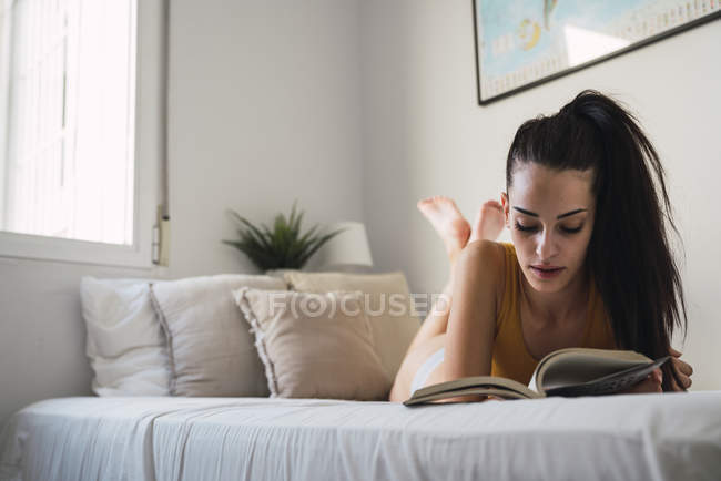 Mujer joven acostada en la cama en casa y leyendo un libro - foto de stock