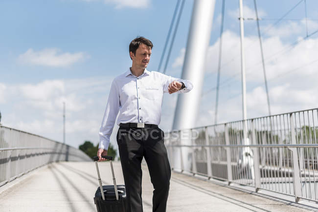 Empresario con maleta enrollable en un puente comprobando la hora — Stock Photo