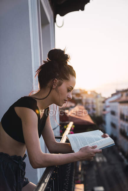 Приваблива молода жінка на балконі над містом на заході читання книг — стокове фото