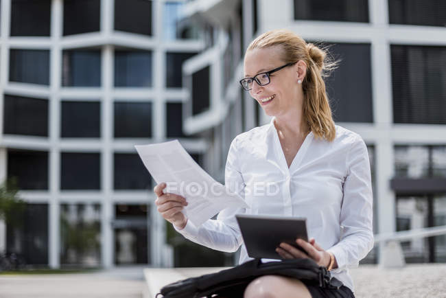 Портрет улыбающейся деловой женщины с документами и планшетом, сидящей перед офисным зданием — стоковое фото