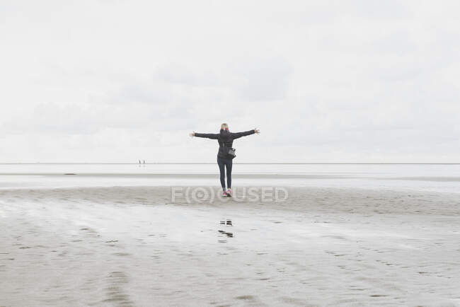 Países Bajos, Ouddorp, vista trasera de la mujer con los brazos extendidos de pie en la playa en otoño - foto de stock