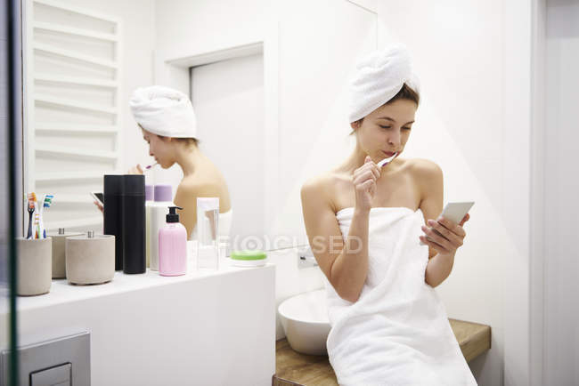 Молода жінка у ванній кімнаті чистити зуби під час читання повідомлень на мобільному телефоні — стокове фото