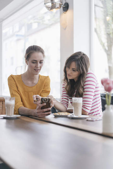 Two girlfriends meeting in a coffee shop, using smartphone — Stock Photo