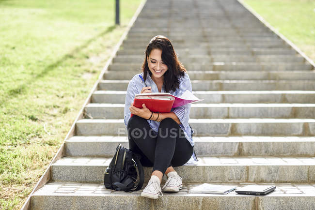 Estudiante sonriente sentado en las escaleras al aire libre tomando notas en un cuaderno - foto de stock