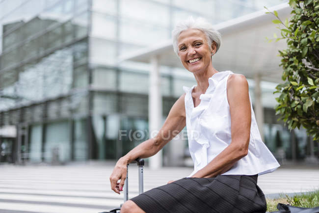 Portrait of happy senior woman with baggage sitting in city — Stock Photo